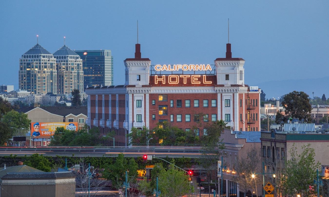 A photo of the front and side façade of a red brick and white stucco building topped by a marquee with a lit sign that reads “California Hotel.” A freeway is in the foreground. A photo of the front and side façade of a red brick and white stucco building topped by a marquee with a lit sign that reads “California Hotel.” A freeway is in the foreground.