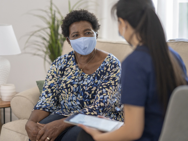 Two women wearing surgical masks sit facing each other on a couch.