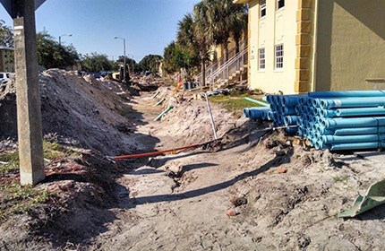 Photograph of a shallow trench running along a street in front of several residential buildings, with plastic pipes stored nearby.