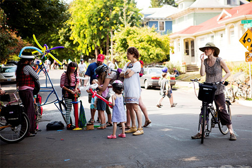 Image of children and adults gathering on a neighborhood street to receive balloon animals. Pedestrians and a bicyclist are also visible in the photograph.