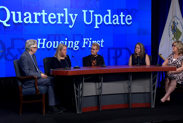 Panelists sit at a table in front of a backdrop that says “Quarterly Update Housing First”. Panelists sit at a table in front of a backdrop that says “Quarterly Update Housing First”.
