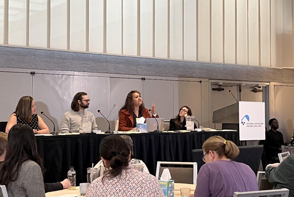 Four panelists sit at a table with the audience in the foreground. Four panelists sit at a table with the audience in the foreground.