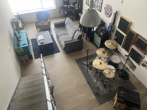 Photograph of apartment loft interior with steps leading down to a living area with a loveseat sofa, chair, and a music area with a drum set and artwork on the walls.