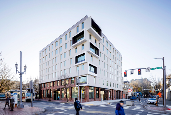 Seven story apartment building at the corner of an intersection with pedestrians, cars, and a light rail train on the street. Seven story apartment building at the corner of an intersection with pedestrians, cars, and a light rail train on the street.