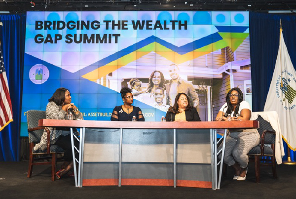 Four panelists sit at a table in front of a backdrop that reads “Bridging the Wealth Gap Summit”.