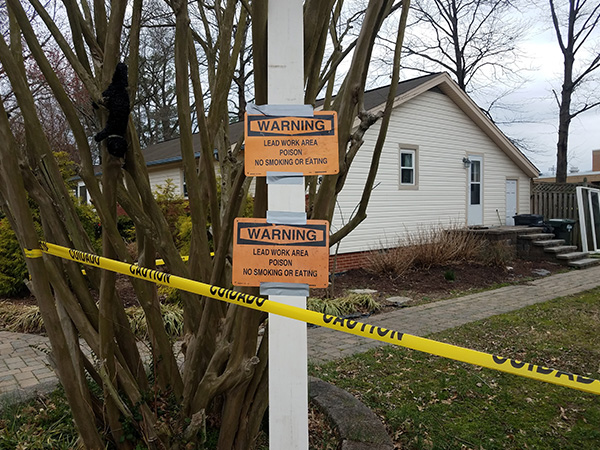 Photo of a house with yellow caution tape blocking it off in the foreground and two orange signs on a post reading “warning lead work area poison no eating or smoking”.