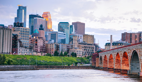 Photograph of downtown Minneapolis, with the Mississippi River and the Stone Arch Bridge in the foreground.